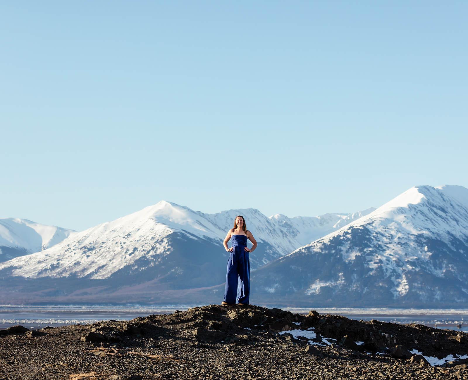 Chelsea Kovalcsik站在外面，双臂叉腰, 在一个阳光明媚的日子里，在美丽的阿拉斯加山脉前，天空湛蓝